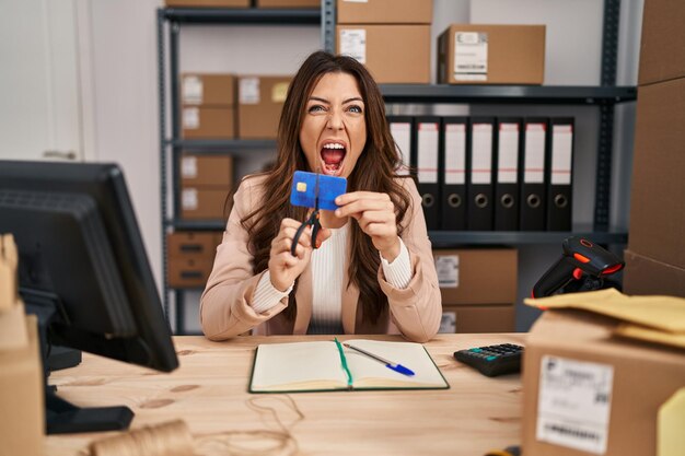 Joven morena que trabaja en el comercio electrónico de pequeñas empresas cortando tarjetas de crédito enojada y enojada gritando frustrada y furiosa, gritando de ira. rabia y concepto agresivo.