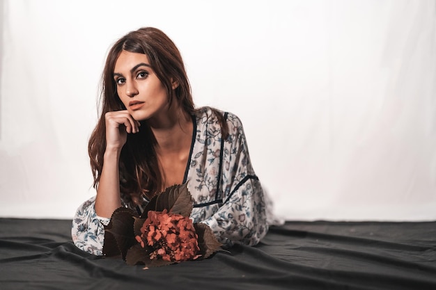 Una joven morena de raza caucásica con un vestido de flores vintage sobre un fondo blanco. Retrato con una flor roja en la mano, acostada sobre un paño negro con una mirada dulce