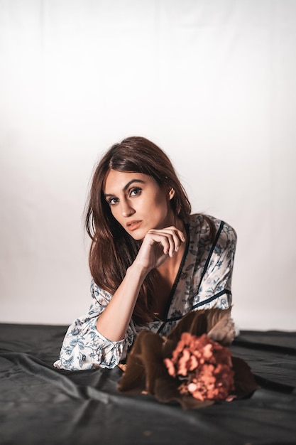 Una joven morena de raza caucásica con un vestido de flores vintage sobre un fondo blanco. Retrato con una flor roja en la mano, acostada sobre un paño negro con una mirada dulce