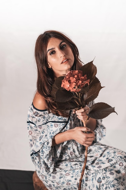 Una joven morena de raza caucásica con un vestido de flores vintage sobre un fondo blanco. Retrato con una flor roja en la mano, con una mirada dulce sentada en el estudio