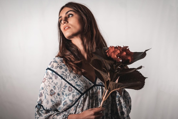 Una joven morena de raza caucásica con un vestido de flores vintage sobre un fondo blanco. Retrato con una flor roja en la mano, con una mirada dulce