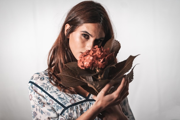 Una joven morena de raza caucásica con un vestido de flores vintage sobre un fondo blanco. Retrato con una flor roja en la mano, con una mirada dulce