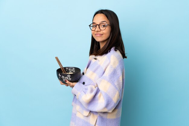 Joven morena de raza mixta mujer sosteniendo un cuenco lleno de fideos sobre la pared azul aislada que planea algo