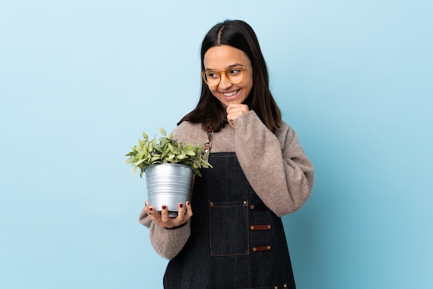 Joven morena de raza mixta mujer sosteniendo una planta sobre pared azul aislado mirando hacia el lado y sonriendo