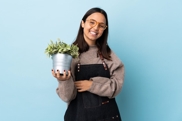 Foto joven morena de raza mixta mujer sosteniendo una planta sobre pared azul aislado riendo