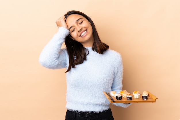 Joven morena de raza mixta mujer sosteniendo sushi sobre pared aislada riendo