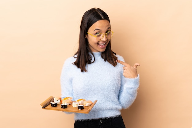 Joven morena de raza mixta mujer sosteniendo sushi sobre la pared dando un gesto de pulgares arriba