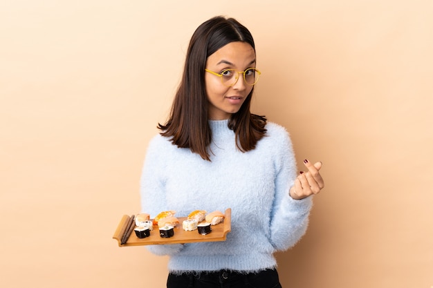 Foto joven morena de raza mixta mujer sosteniendo sushi sobre pared haciendo gesto de dinero