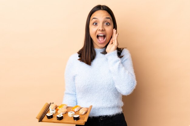 Joven morena de raza mixta mujer sosteniendo sushi sobre la pared con sorpresa y expresión facial conmocionada