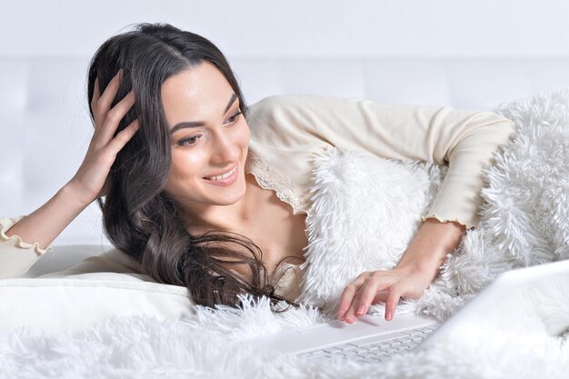 joven morena relajándose en la cama, usando una laptop