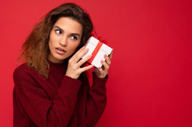 joven morena rizada mujer aislada sobre fondo rojo pared vistiendo un suéter rojo con caja de regalo mirando hacia un lado y pensando.