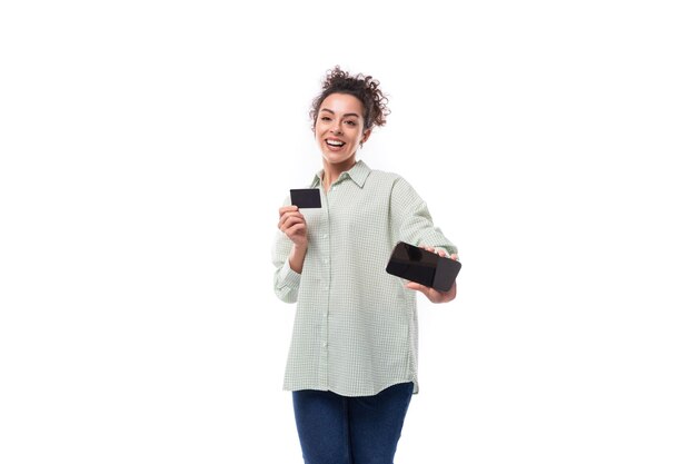 Foto una joven morena rizada sonriente vestida con una camisa a cuadros que sostiene un teléfono inteligente de tarjeta de crédito