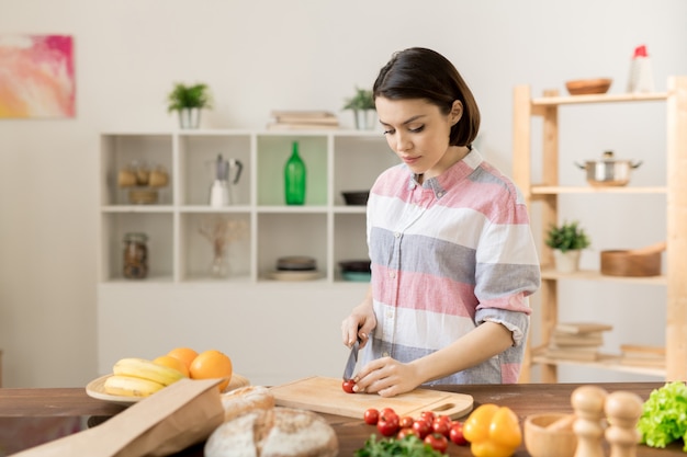 Joven morena en ropa casual de pie junto a la mesa de la cocina y cortando tomates frescos para ensalada