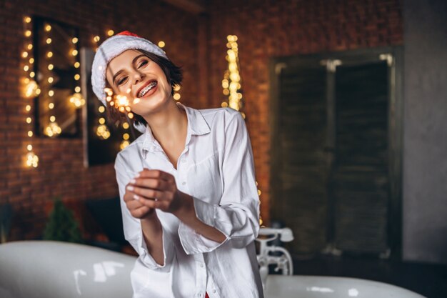 Una joven morena sentada en el borde del baño con sombrero de navidad sosteniendo la luz de Bengala de fuegos artificiales de bengala ardiente en las manos