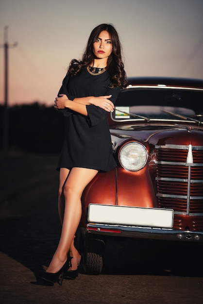 Una joven morena sexy con piernas largas con un vestido negro y zapatos de tacón se encuentra cerca de un coche retro burdeos en una carretera rural a última hora de la noche