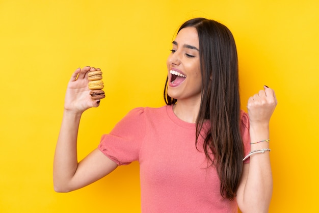 Joven morena sobre amarillo con coloridos macarons y celebrando una victoria