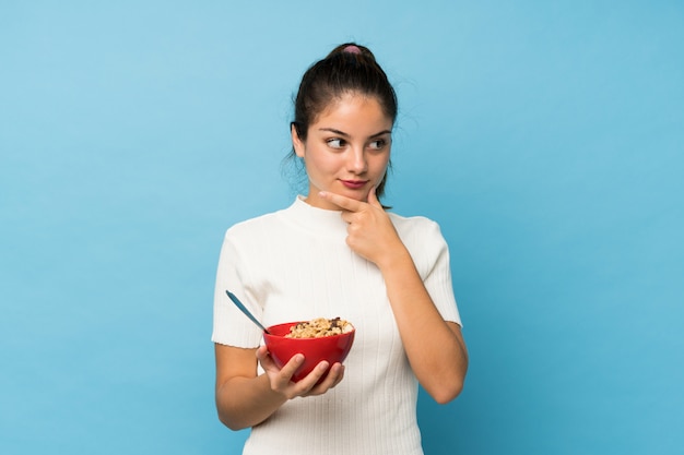 Joven morena sobre azul aislado sosteniendo un tazón de cereales y pensando