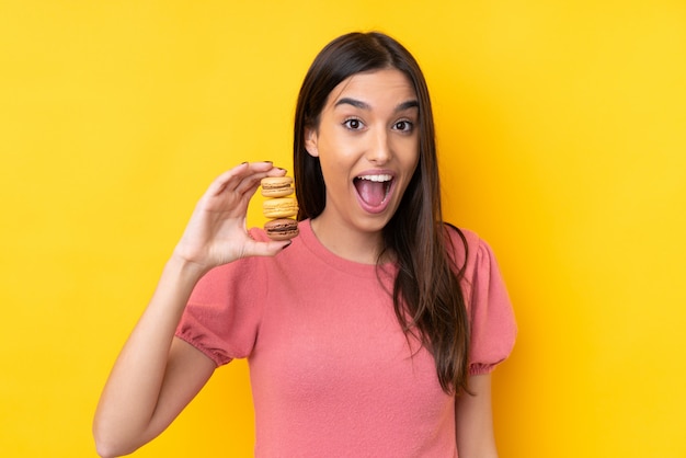 Joven morena sobre pared amarilla aislada con coloridos macarons franceses con expresión de sorpresa