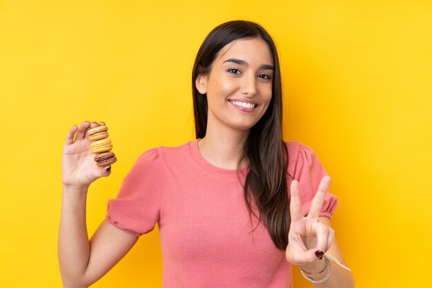 Joven morena sobre pared amarilla aislada sosteniendo coloridos macarons franceses y mostrando el signo de la victoria