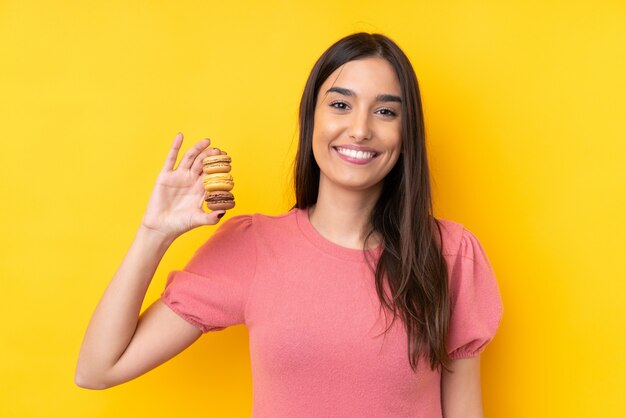 Joven morena sobre pared amarilla aislada sosteniendo coloridos macarons franceses y sonriendo mucho