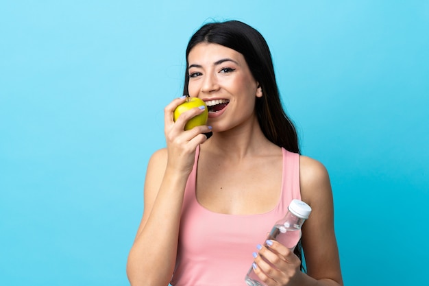 Joven morena sobre pared azul aislada con una botella de agua y comer una manzana