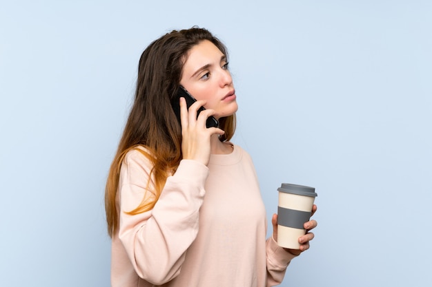 Joven morena sobre pared azul aislada con café para llevar y un móvil