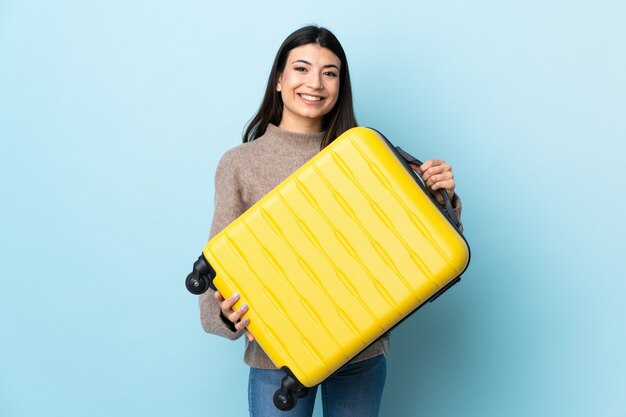 Joven morena sobre pared azul aislada en vacaciones con maleta de viaje