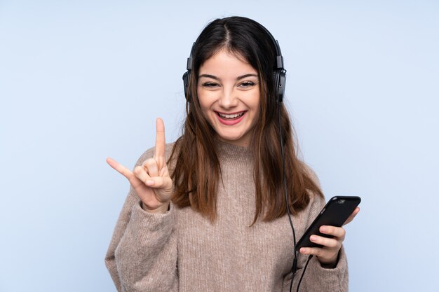 Joven morena sobre pared azul escuchando música con un móvil haciendo gesto de rock