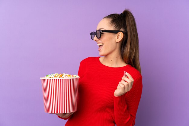 Joven morena sobre pared púrpura aislada con gafas 3d y sosteniendo un gran cubo de palomitas de maíz mientras mira hacia el lado