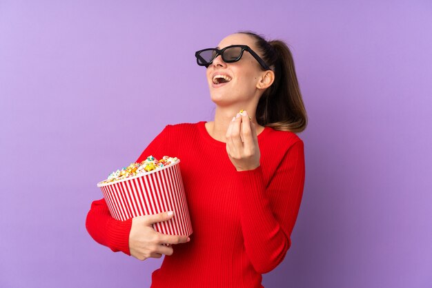 Joven morena sobre pared púrpura aislada con gafas 3d y sosteniendo un gran cubo de palomitas de maíz