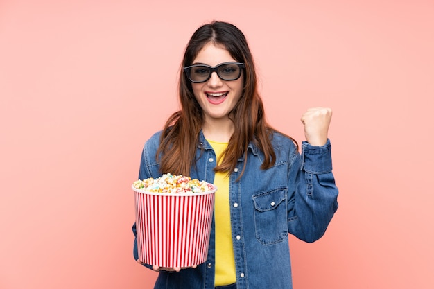 Joven morena sobre pared rosa con gafas 3d y sosteniendo un gran cubo de palomitas de maíz