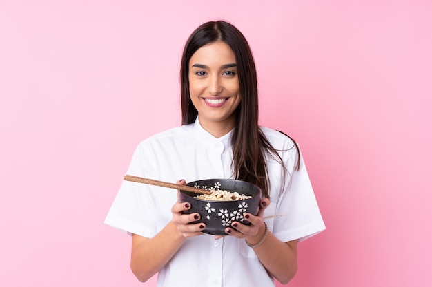 Joven morena sobre rosa sonriendo mucho mientras sostiene un tazón de fideos con palillos