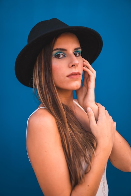 Una joven morena con un sombrero negro y una camiseta blanca de fondo azul