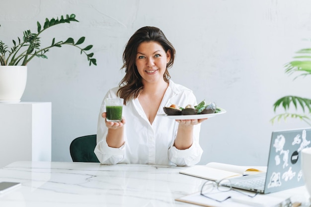 Joven morena sonriente doctora nutricionista de talla grande con camisa blanca con comida verde saludable en la sala de oficina moderna y luminosa El doctor se comunica con el paciente en línea