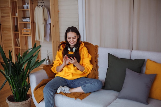 Una joven morena sonriente con el pelo largo en una sudadera amarilla brillante, jeans azules y zapatillas blancas se sienta en casa en un sofá gris con un teléfono inteligente en las manos, mira la pantalla
