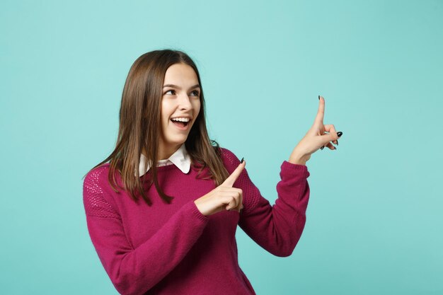 Una joven morena sonriente con ropa informal posando aislada en un retrato de estudio de fondo de pared verde azul. Gente emociones sinceras concepto de estilo de vida. Burlarse de señalar con la mano del dedo en el espacio de la copia