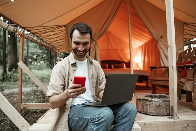 Joven morena sonriente sentado en glamping con ordenador portátil y smartphone, streaming, mensajería. Estilo de vida de camping. Viajes de bajo presupuesto. Concepto de trabajo remoto. Tecnología moderna de comunicación por internet.