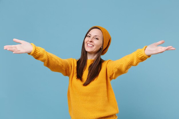 Una joven morena sonriente con un sombrero de suéter amarillo posando aislada en un retrato de estudio de fondo azul. Gente emociones sinceras concepto de estilo de vida. Simulacros de espacio de copia. De pie con las manos extendidas.