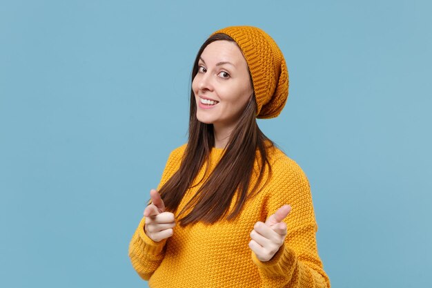 Una joven morena sonriente con un sombrero de suéter amarillo posando aislada en un retrato de estudio de fondo azul. Gente emociones sinceras concepto de estilo de vida. Simulacros de espacio de copia. Señalando con el dedo índice a la cámara.