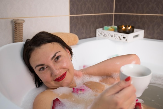 Joven morena sonriente tomando un baño con pétalos de rosa en casa haciendo tratamientos de spa para el equilibrio y la salud mental