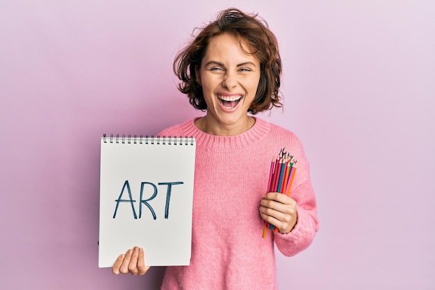 Joven morena sosteniendo un cuaderno de arte y lápices de colores sonriendo y riéndose a carcajadas porque es una broma divertida y loca