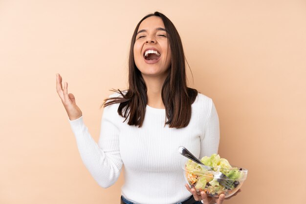 Joven morena sosteniendo una ensalada sobre pared aislada sonriendo mucho