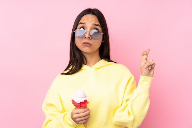 Joven morena sosteniendo un helado de cucurucho sobre pared rosa aislado con dedos cruzando y deseando lo mejor