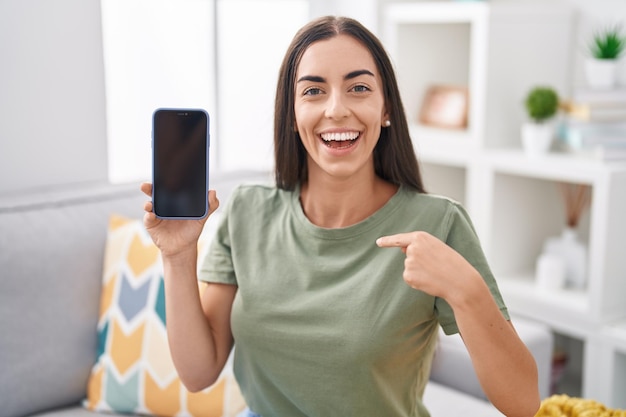 Joven morena sosteniendo un teléfono inteligente mostrando una pantalla en blanco apuntando con el dedo a uno mismo sonriendo feliz y orgulloso