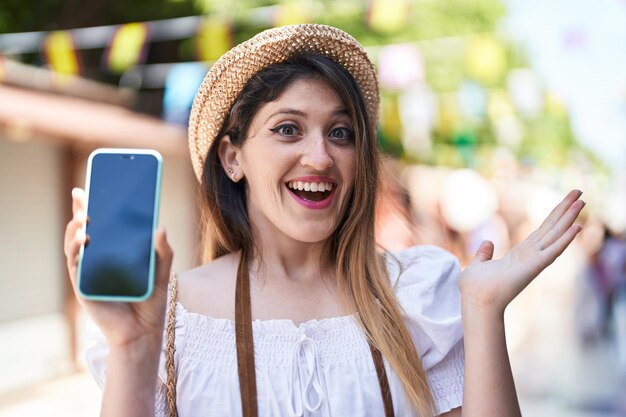 Joven morena sosteniendo un teléfono inteligente que muestra una pantalla en blanco celebrando el logro con una sonrisa feliz y una expresión ganadora con la mano levantada