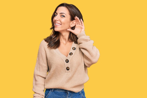 Joven morena con suéter casual de invierno sonriendo con la mano sobre la oreja escuchando rumores o chismes. concepto de sordera.