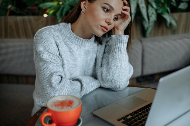 Una joven morena con un suéter gris trabaja en una computadora portátil en un café Se ve frustrada con la computadora portátil