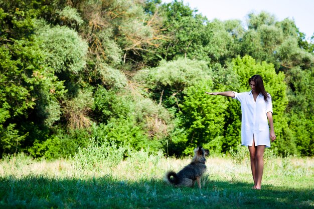 Joven morena con vestido blanco jugando con perro en el césped en día de verano con árboles verdes y río