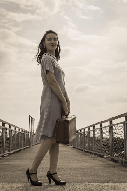 Una joven morena con un vestido y una maleta vintage en las manos se levanta contra un cielo azul claro Estilo retro vintage Cielo pacífico