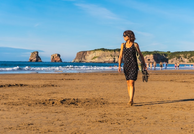 Una joven morena con un vestido negro y gafas de sol caminando por la playa en Hendaya, País Vasco francés. Francia. Paseando una tarde de verano